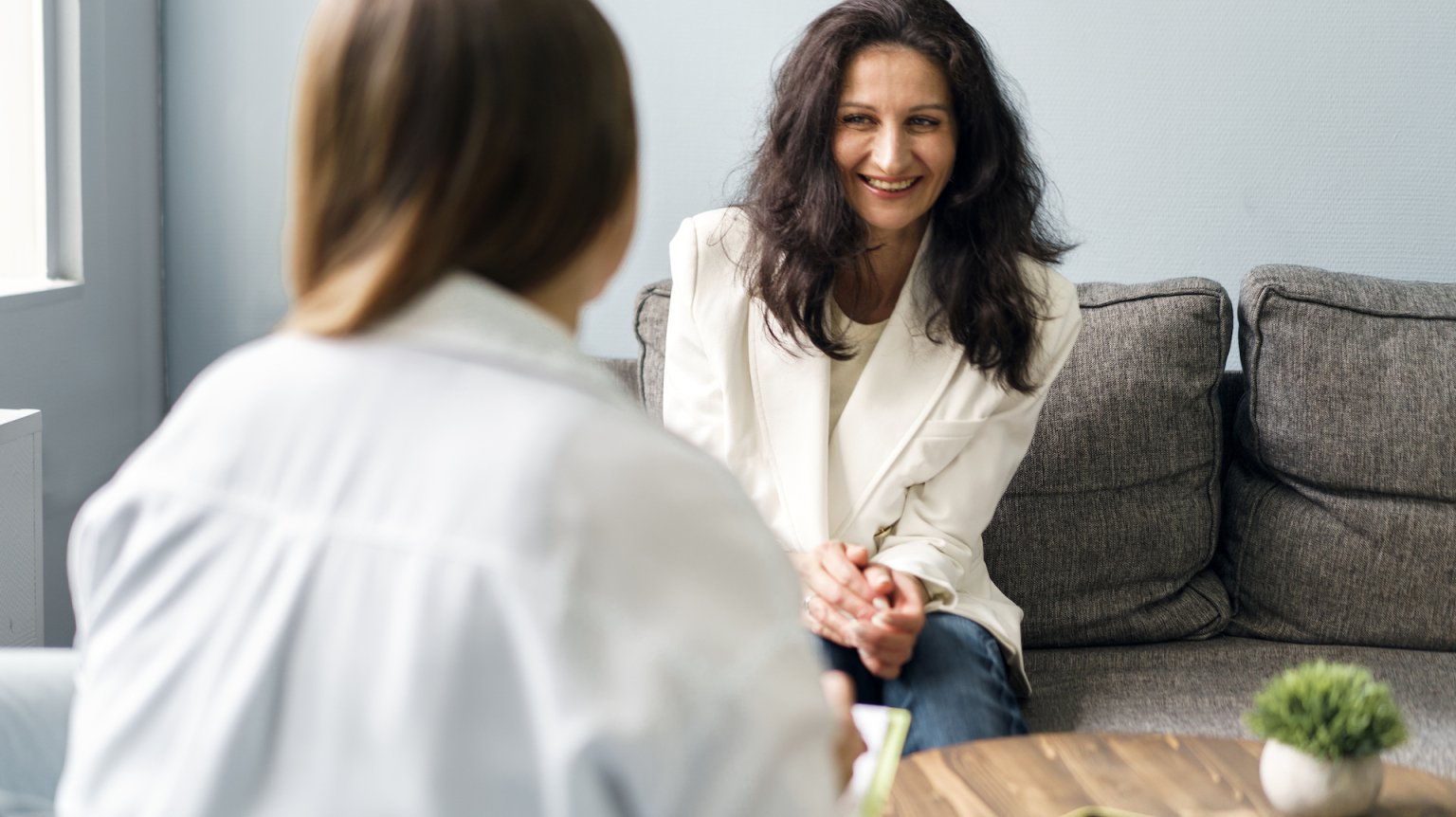 A female therapist talks to a woman sitting on a couch. A phone is on the table. 