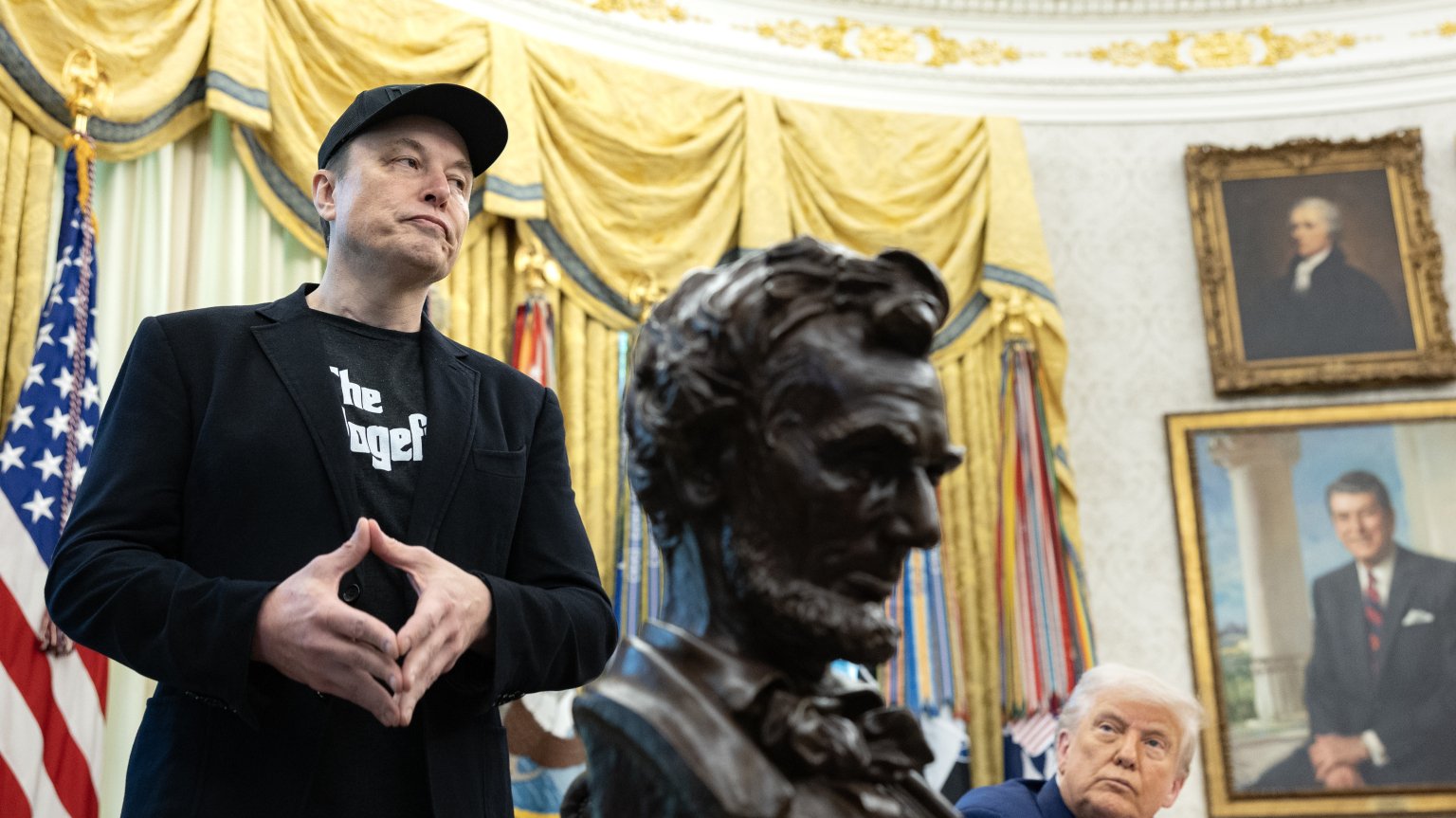 Elon Musk stands in the oval office next to Trump, who stares up at him. Between them is a bust of Abraham Lincoln.