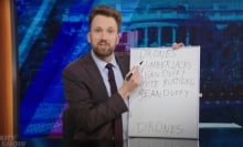 A man in a suit behind a talk show desk points a pen at a small whiteboard covered in writing.