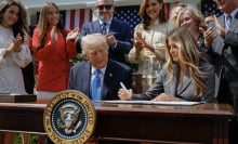 President Donald Trump and Melania Trump sit at a desk and sign the Take It Down Act while surrounded by people clapping.