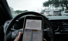 A hand holding an iPhone in front of a steering wheel. The phone shows an emergency alert notification for a storm surge.