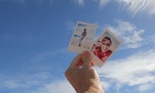 One hand holding up two instant photos of friends against blue sky