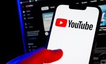 A hand holds a smartphone showing the YouTube logo. A computer screen glows in a darkened room behind it.