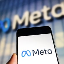 A phone displaying the Meta logo held up in front of a Meta office building.