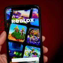A hand holds a phone showing the Roblox homepage.