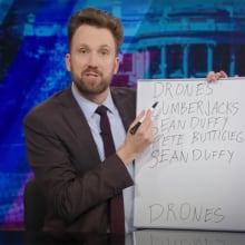 A man in a suit behind a talk show desk points a pen at a small whiteboard covered in writing.