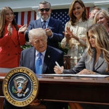 President Donald Trump and Melania Trump sit at a desk and sign the Take It Down Act while surrounded by people clapping.