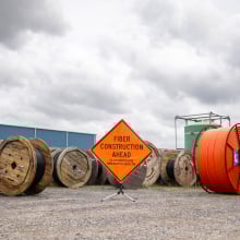 A group of large spools of orange and black fiber optic cables on a construction site. An orange sign reads, "Fiber construction ahead."