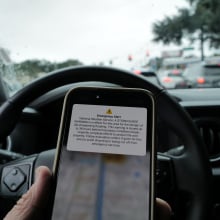 A hand holding an iPhone in front of a steering wheel. The phone shows an emergency alert notification for a storm surge.