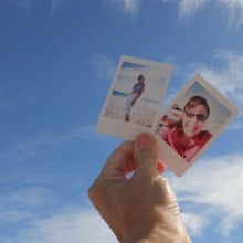 One hand holding up two instant photos of friends against blue sky