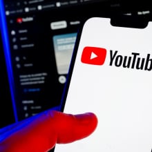 A hand holds a smartphone showing the YouTube logo. A computer screen glows in a darkened room behind it.