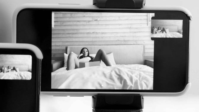 An iPhone sits on a tripod, filming a woman who lays with her legs open on a bed.