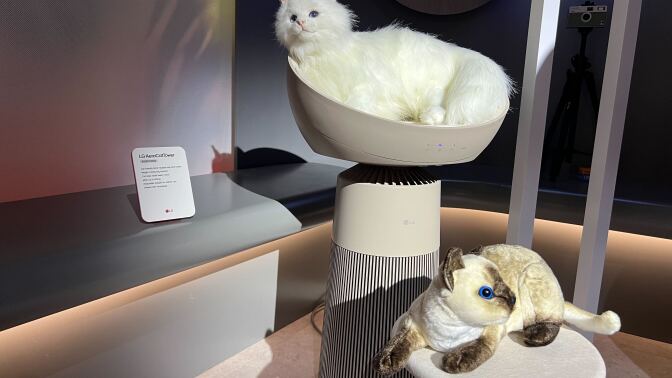 A display featuring the LG AeroCatTower, with a lifelike white cat model resting on the tower's top section and a plush Siamese cat on a smaller platform.