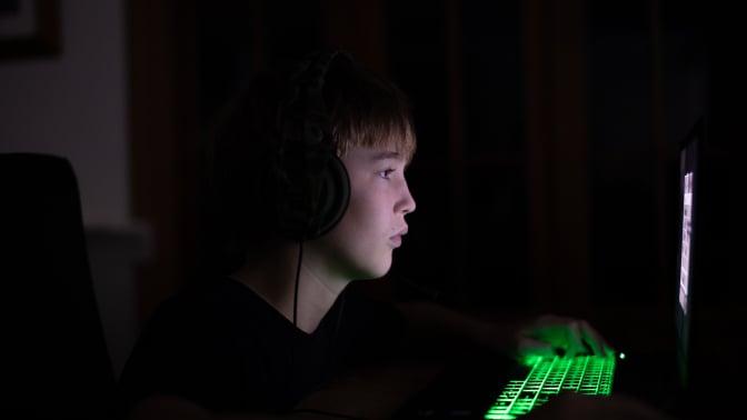 A tween or teen boy looks at a computer screen in a darkened room. 