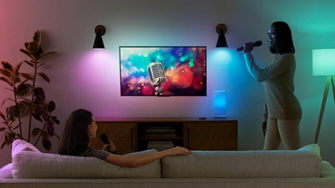 two people are in a dimly lit room aside from two amazon smart bulbs that are displaying purple and blue colors. One person is sitting on the couch and the other is standing while singing into a microphone