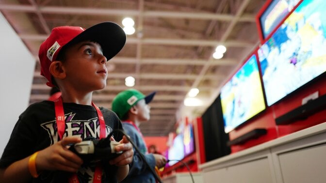 Two small children wearing Nintendo merch play a new Switch 2 game in front of large monitors. 