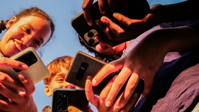 A group of young kids smile and hold out several different models of phones while huddled in a group.