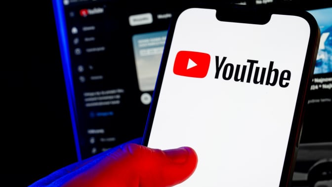 A hand holds a smartphone showing the YouTube logo. A computer screen glows in a darkened room behind it.