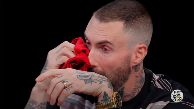 A man sits in a dark studio dabbing his eyes with a red napkin.