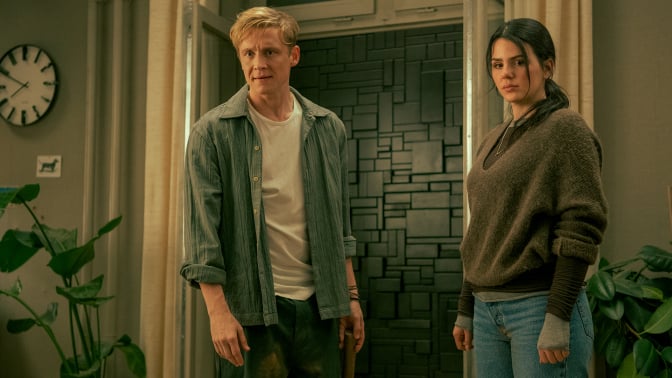 A man and a woman stand in a room with a black wall covering a doorway in the background.
