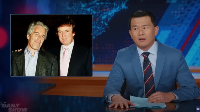 A man in a suit sits behind a talk show desk, side-eyeing another image of two men standing next to each other.