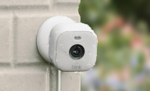 A white Blink Mini 2 security camera mounted on a light-colored outdoor wall, covered in water droplets.