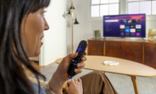 woman speaking into Roku voice remote and watching tv through Roku Streaming Stick Plus