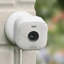A white Blink Mini 2 security camera mounted on a light-colored outdoor wall, covered in water droplets.