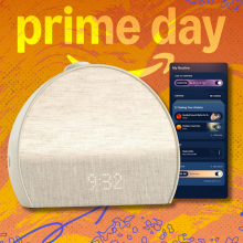 A photo composite of the Hatch Restore 3 alarm clock in a putty color, shown against a stylized orange and yellow Prime Day background. A smartphone displaying the Hatch app is visible next to the clock.