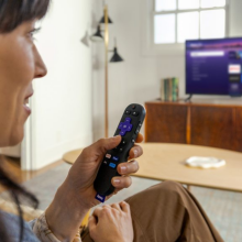 woman speaking into Roku voice remote and watching tv through Roku Streaming Stick Plus