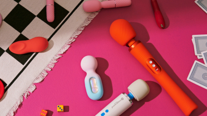 Flat lay of assorted colorful sex toys, including wand and bullet vibrators, scattered with dice and cards on a bright pink surface and textured white mat.